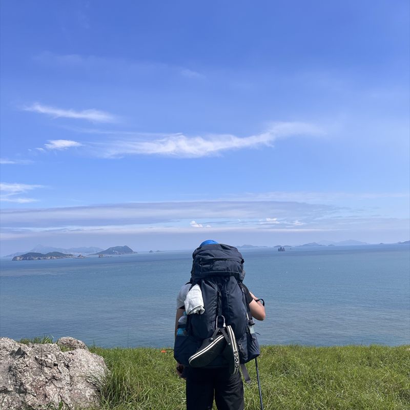 A backpacker standing on a grass headland, facing islands across open water